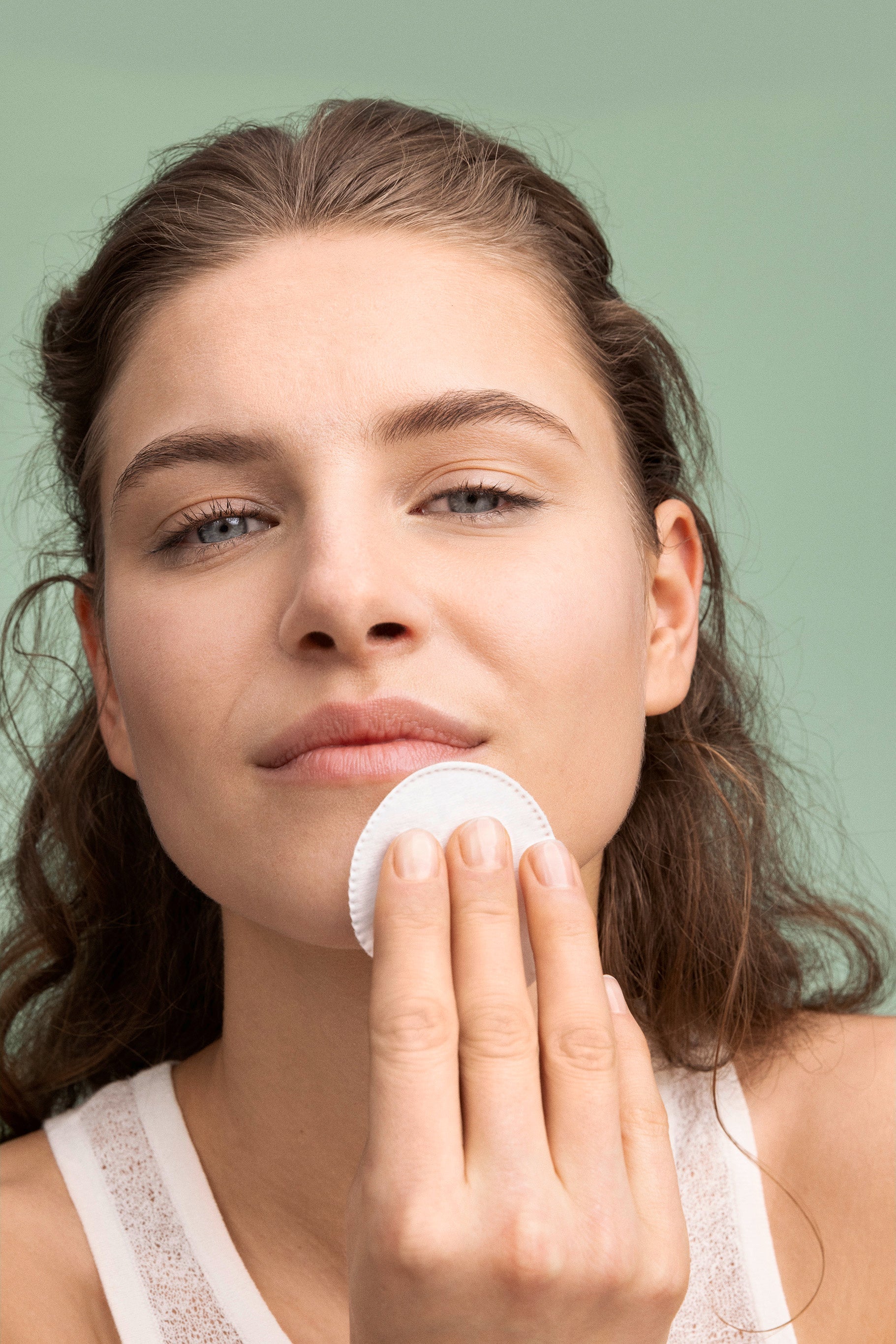 Junge Frau reinigt ihr Gesicht sanft mit einem Wattepad vor grünem Hintergrund.