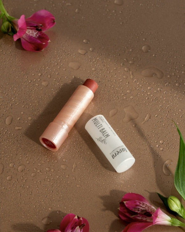 lavera Multibalm mit roséfarbenem Inhalt liegt auf einer taufeuchten braunen Oberfläche, umgeben von pinken Blüten.