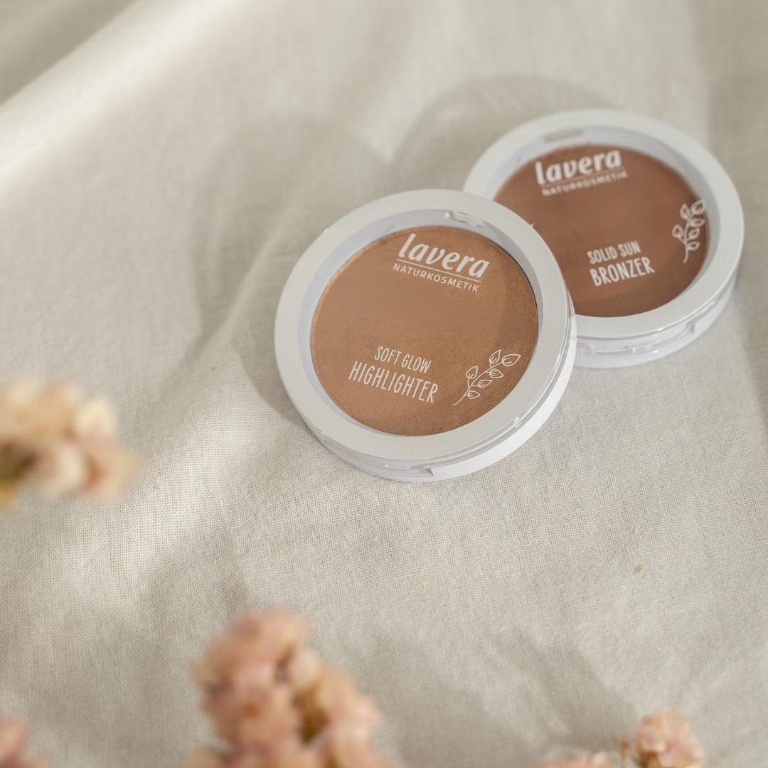lavera Teintprodukte, ein Soft Glow Highlighter und ein Solid Sun Bronzer, liegen auf hellem Leinenstoff neben unscharfen rosafarbenen Blüten.