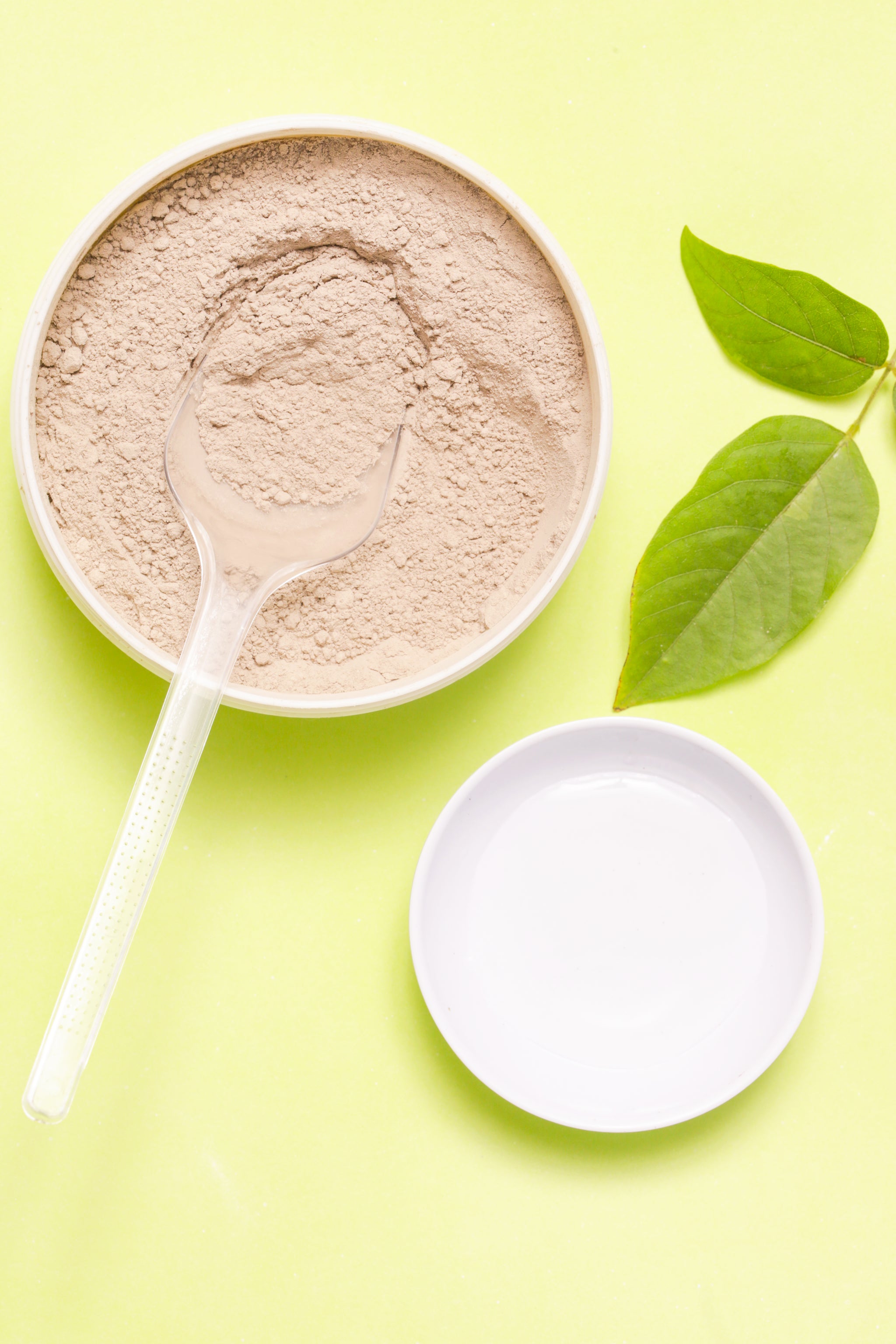 Schale mit beigefarbener Mineralerde und einem durchsichtigen Löffel, daneben ein weißer Teller und zwei grüne Blätter auf gelbem Hintergrund.