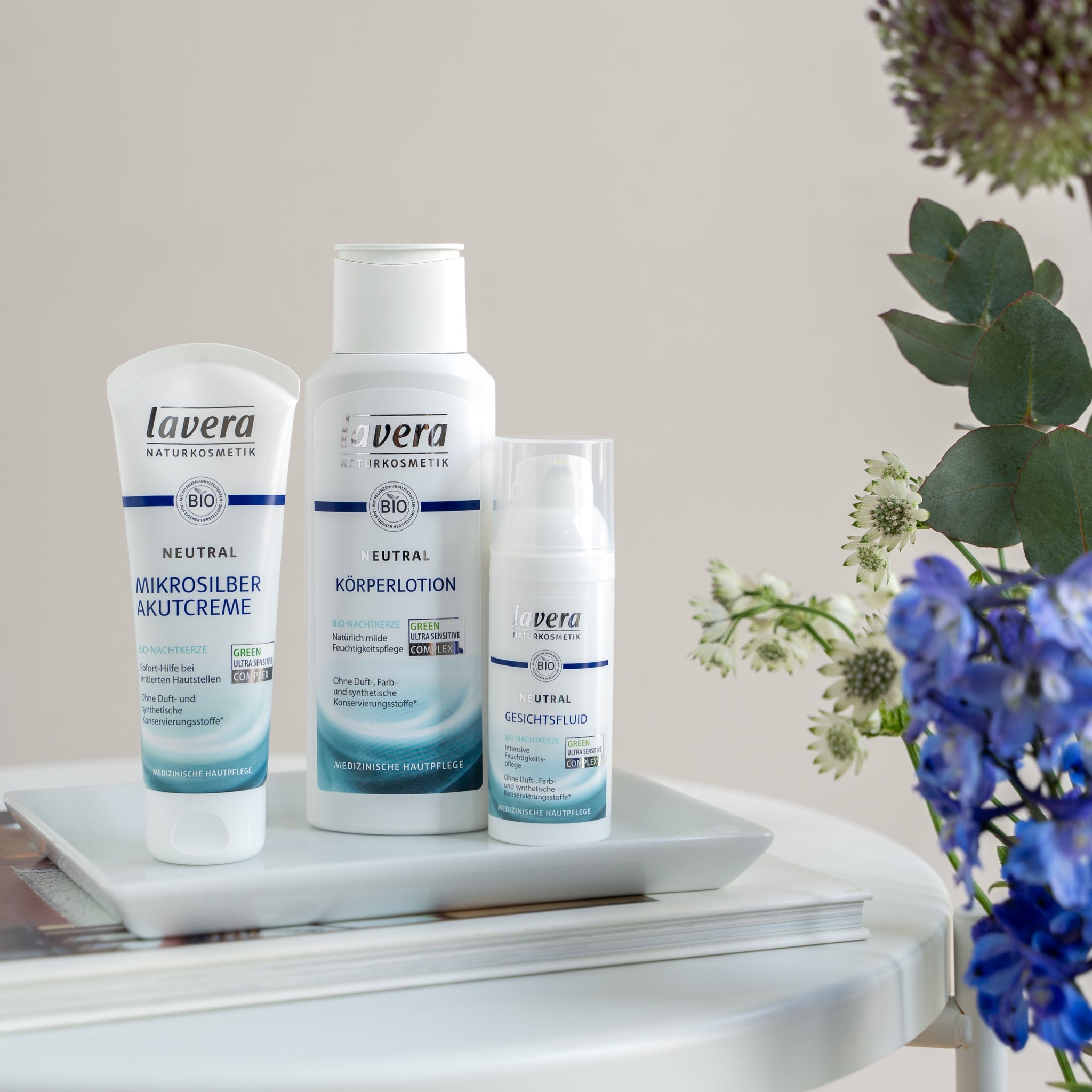 Produits de soin cutané médical lavera Neutral avec microargent : crème SOS, lotion corps et fluide visage, disposés de manière décorative avec des fleurs sur une table blanche.