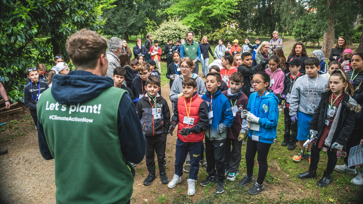 Junger Mann mit grüner Weste steht vor einer Gruppe von Kindern und erzählt - draußen im Grünen
