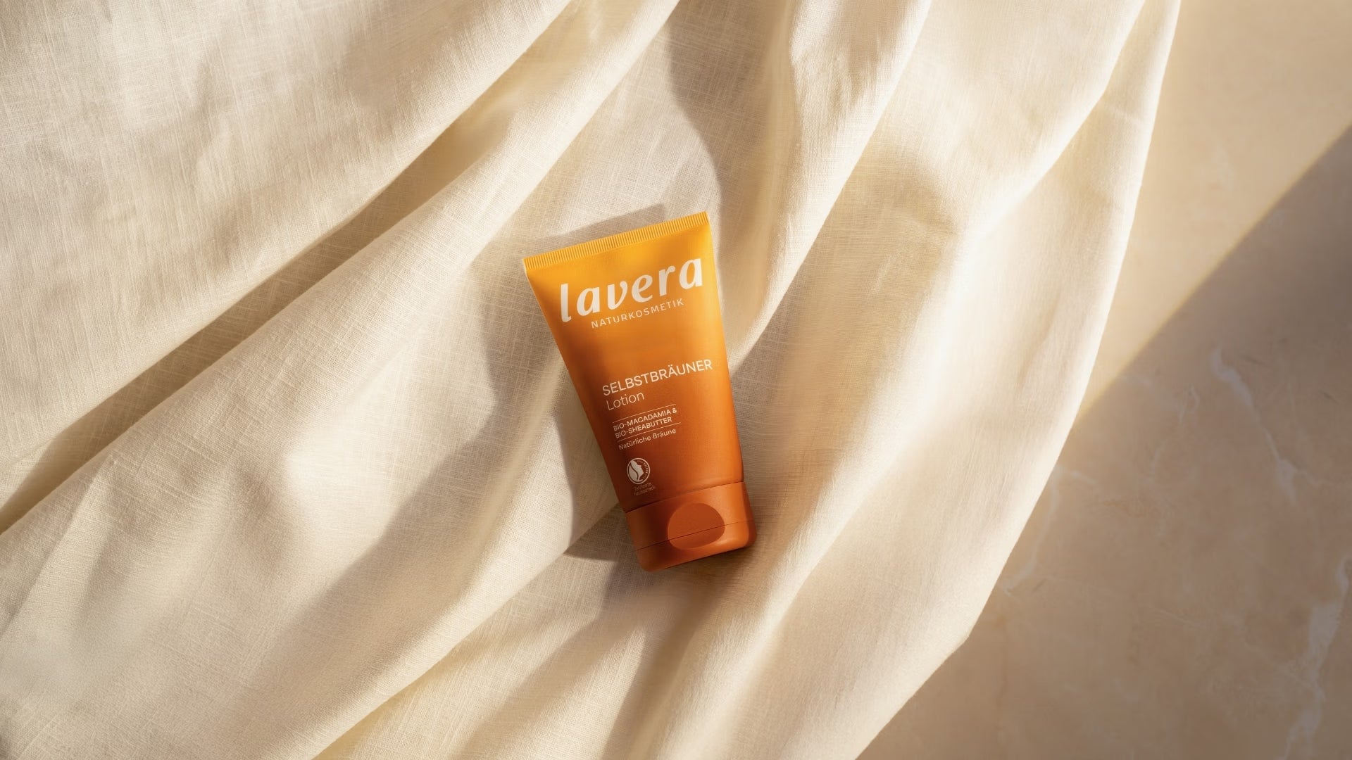 L’autobronzant lavera est posé en biais sur un tissu clair aux drapés souples et baigné d’une lumière solaire chaleureuse.