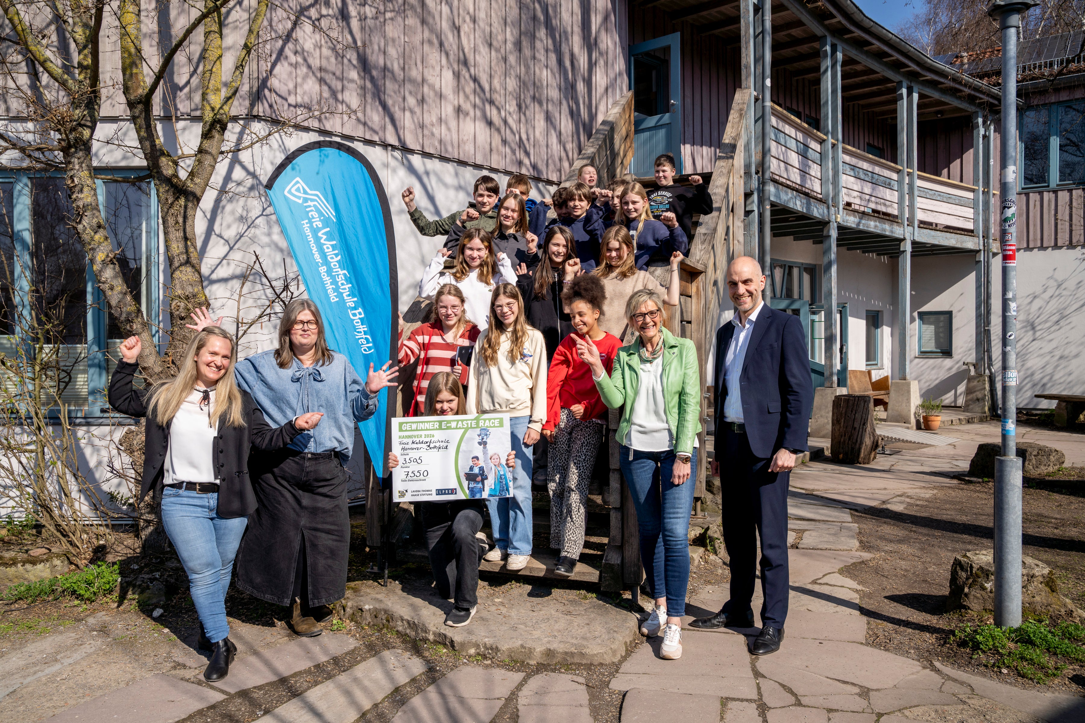 Schulklasse steht auf einer Holztreppe vor einer Schule und hält E-Waste-Race Urkunde in der Hand