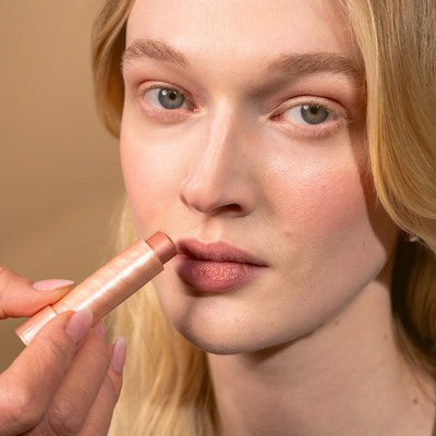 Jeune femme aux cheveux blonds qui sourit à la caméra et s'applique du Multi Balm sur les lèvres