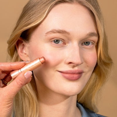 Jeune femme aux cheveux blonds qui sourit à la caméra et s'applique de la couleur sur la joue