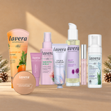 lavera XXL Christmas Set: Acht Produkte nebeneinander auf warmbeigem Hintergrund mit dezenten winterlichen Akzenten: Duschgel Energize Me, Plumping Lip Balm, Depuff & Hydration Eye Patches, Leave-in Spray Smooth & Silk, Calm & Comfort Rich Cream, Beauty Körperöl, High Lash Mascara und basis sensitive Reinigungsschaum.