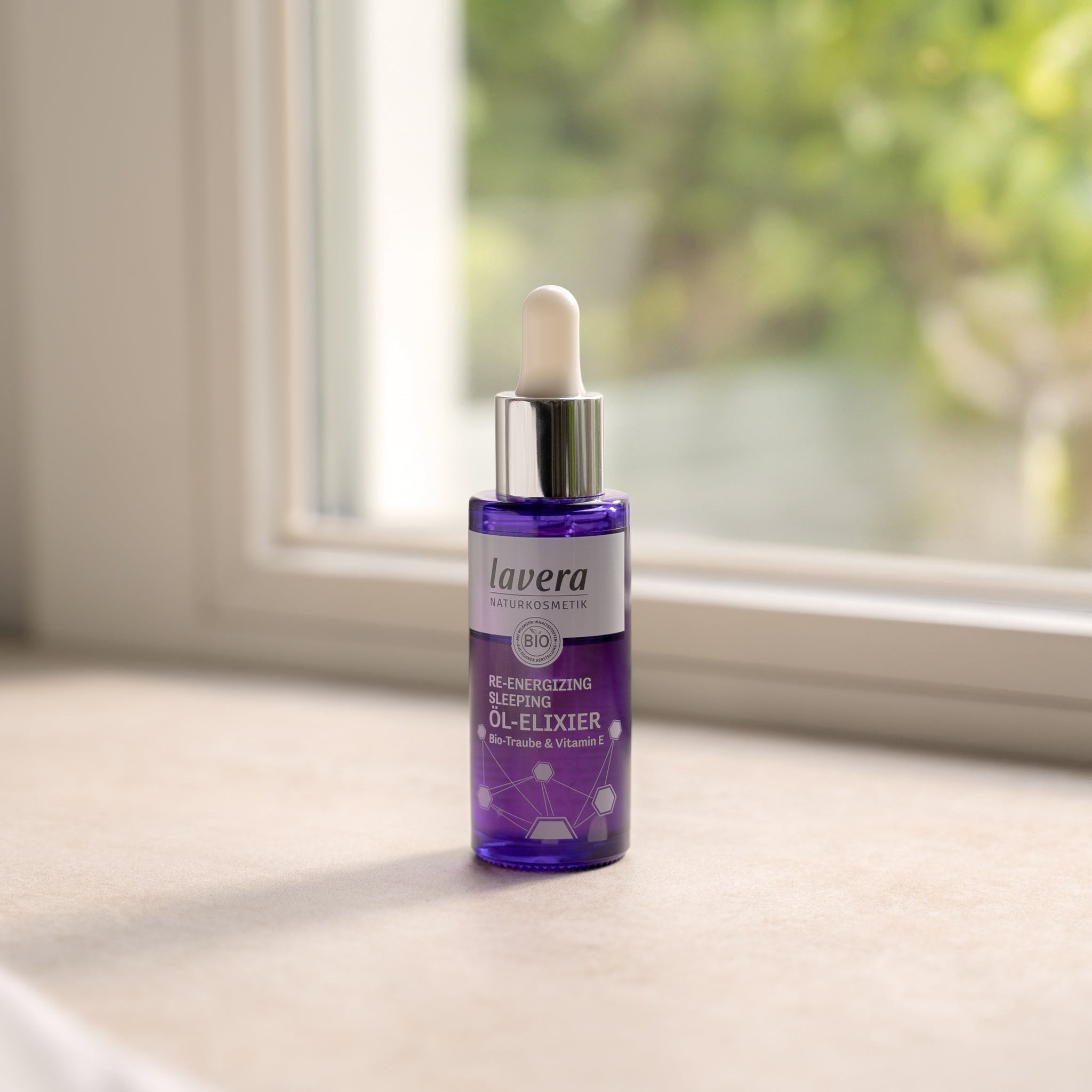 Eine violette Glasflasche mit Pipette des lavera Naturkosmetik Re-Energizing Sleeping Öl-Elixiers  stehend auf einer Fensterbank im weichen Tageslicht, mit verschwommenem Grün im Hintergrund.
