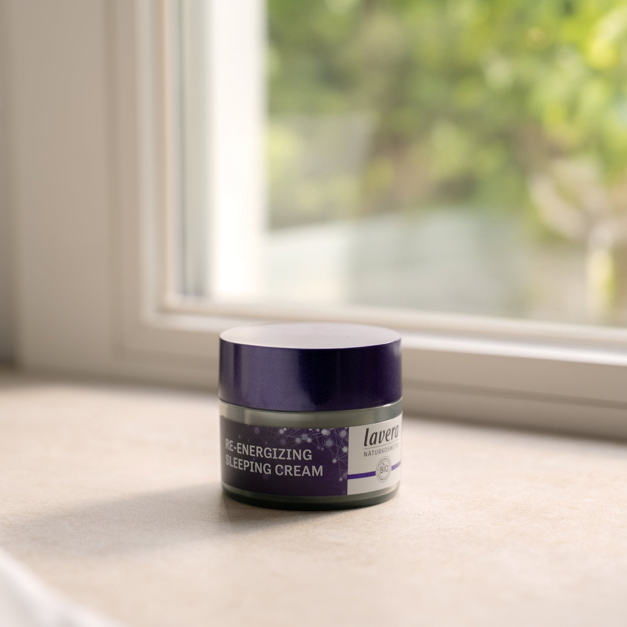 Ein runder Tiegel mit violettem Deckel des lavera Naturkosmetik Re-Energizing Sleeping Cream Produkts, auf einer Fensterbank im weichen Tageslicht stehend, mit verschwommenem grünen Hintergrund.